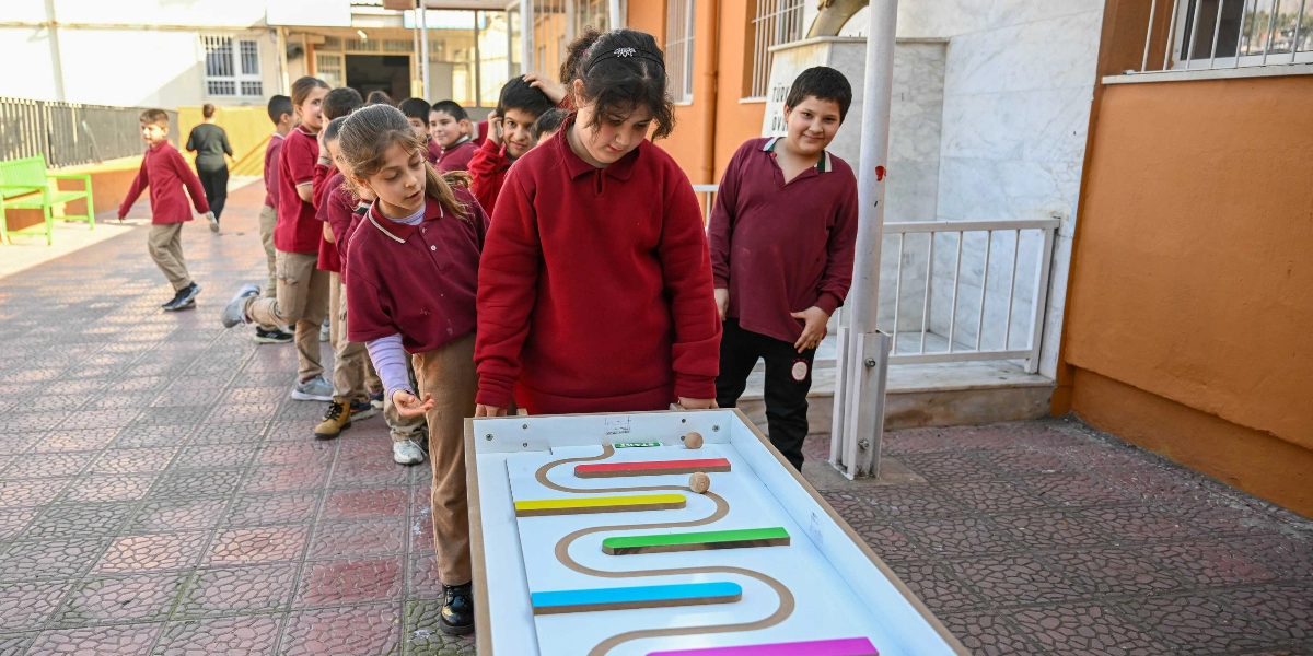 Okul bahçesinde bordo renkli okul formaları giymiş bir grup öğrenci, üzerinde renkli engeller ve ahşap topların bulunduğu bir labirent oyununu sırayla oynuyor. Ön planda bir kız öğrenci oyuna odaklanmışken, arkasında diğer öğrenciler sırayla bekliyor, arka planda ise bahçede koşan başka çocuklar görülüyor.