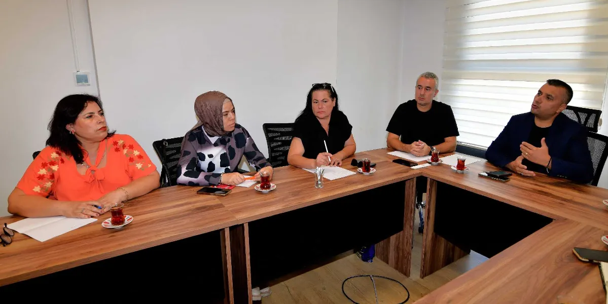 U-şeklindeki bir toplantı masasında oturan beş kişi, bir konu üzerinde görüşme yapıyor. Masanın başında lacivert ceketli bir erkek konuşurken, diğer üç kadın ve bir erkek katılımcı onu dinliyor. Katılımcıların önünde not kağıtları, kalemler ve ince belli bardaklarda çaylar bulunuyor. Toplantı, beyaz duvarlı ve perdeli bir ofis ortamında gerçekleşiyor.