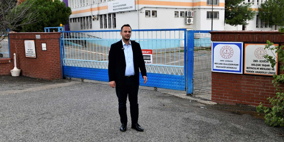 Koyu renk ceket ve açık renk gömlek giymiş bir erkek, İzmir Bornova'daki Selçuk Yaşar Boyacılık Mesleki ve Teknik Anadolu Lisesi ile Mevlana Celaleddin Rumi İmam Hatip Ortaokulu'nun giriş kapısı önünde duruyor. Arka planda okul binası ve üzerinde 