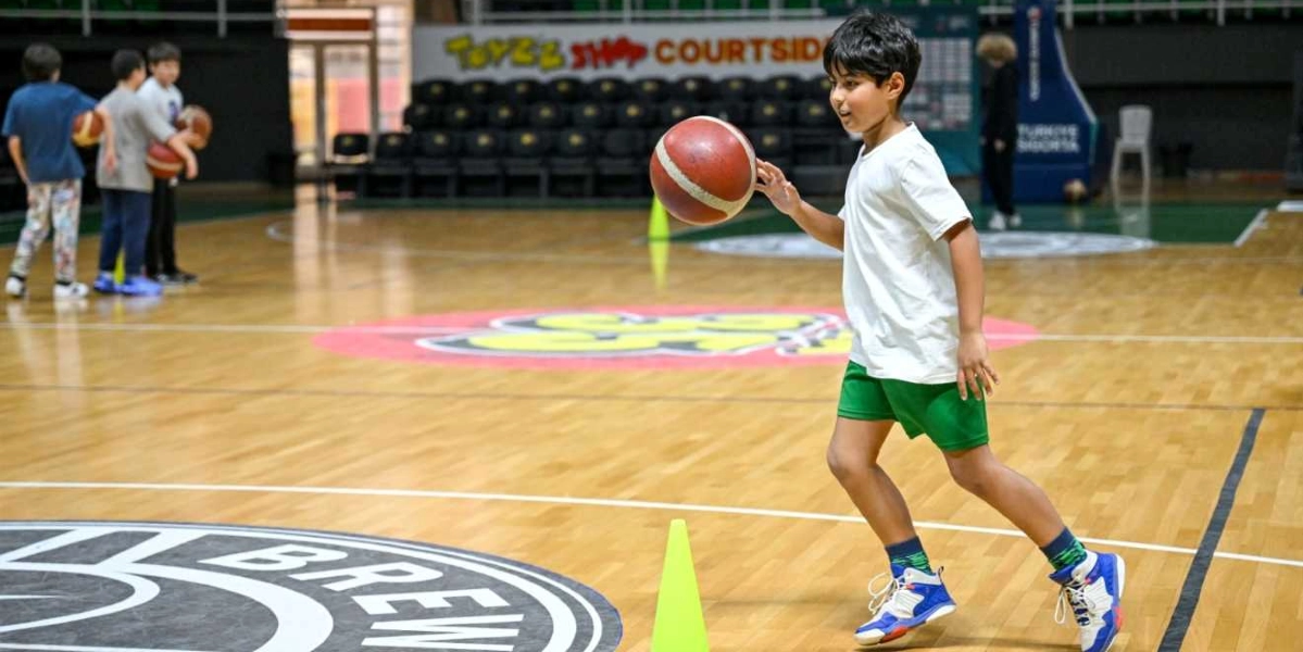 Kapalı bir basketbol sahasında beyaz tişört ve yeşil şort giyen genç bir çocuk, elindeki basketbol topunu sektirerek sarı renkli bir antrenman konisinin yanından geçiyor. Arka planda diğer çocuklar basketbol toplarıyla antrenman yapmaya devam ediyor, saha zemininde ise çeşitli logolar ve yazılar yer alıyor.