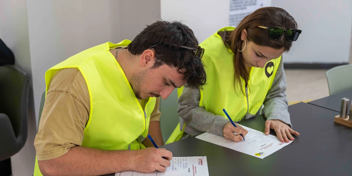 Üzerlerinde neon sarı renkli yelekler bulunan genç bir kadın ve bir erkek, bir masanın başında oturmuş dikkatle form dolduruyor. Genç kadının başında güneş gözlüğü takılı ve yeleğinin üzerinde kalp şeklinde bir logo bulunuyor. Masanın üzerindeki formlarda Bornova Belediyesi'nin kurumsal logoları ve 