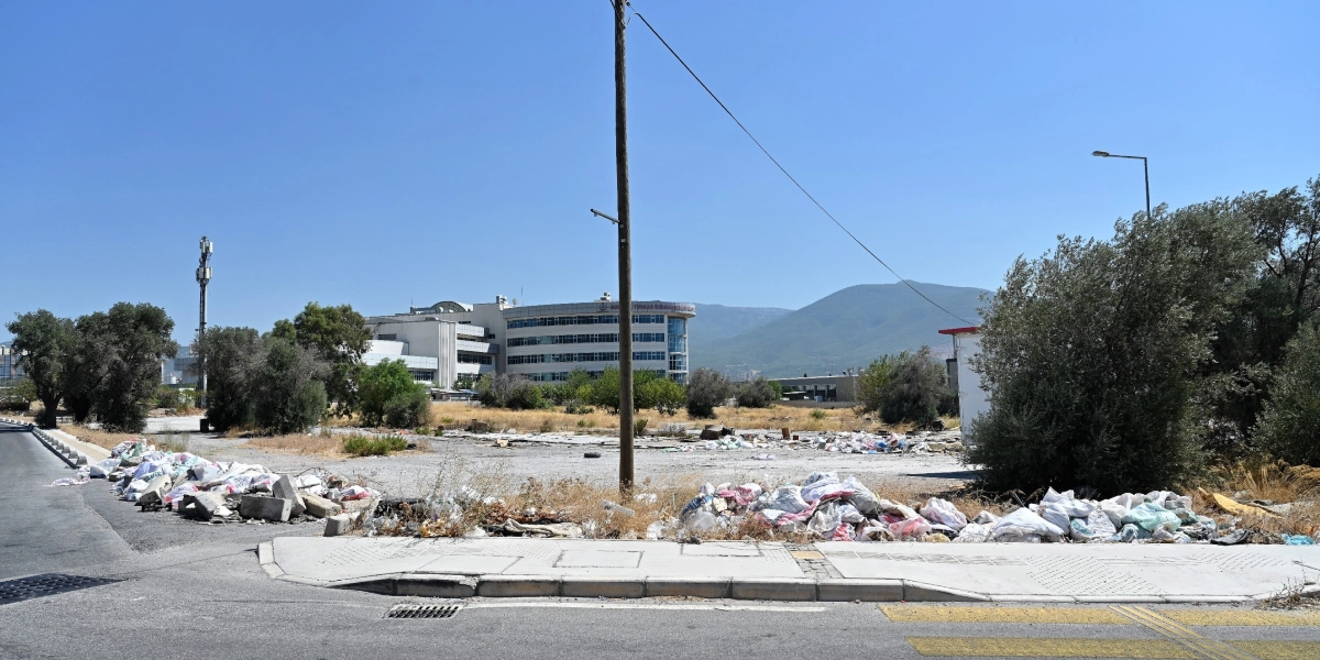 Asfalt bir yolun kenarındaki boş arazide, kaldırım boyunca yığılmış çok sayıda çöp poşeti, inşaat atığı ve moloz yığınları görülüyor. Arka planda ise ağaçlar, geniş bir bina kompleksi ve uzakta bir dağ manzarası yer alıyor.