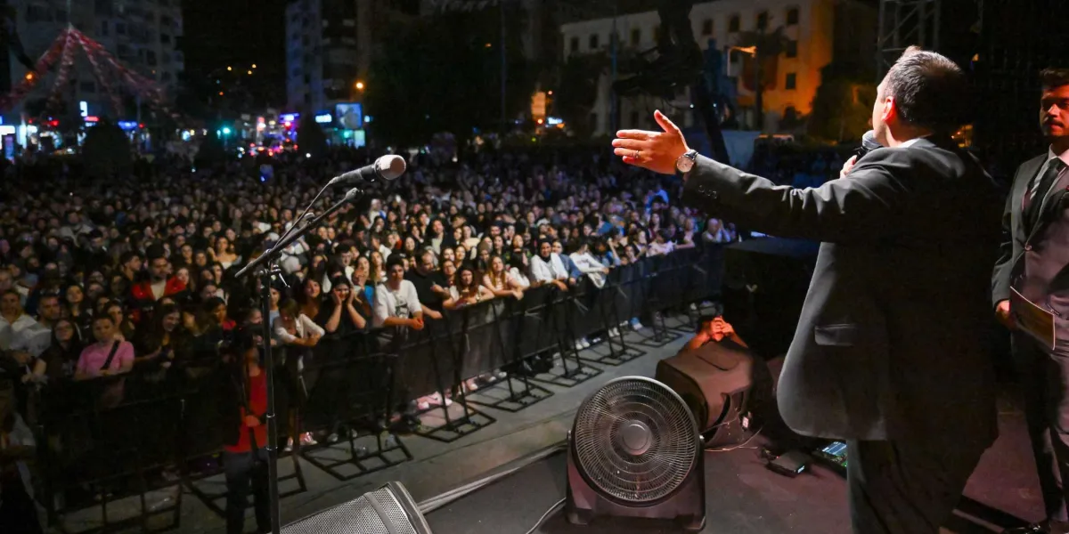 Gece vakti açık havada düzenlenen bir etkinlikte, takım elbiseli bir konuşmacı sahneden kalabalığa hitap ediyor. Konuşmacı sağ elini ileriye doğru uzatarak izleyicilere seslenirken, yanında takım elbiseli başka bir kişi daha duruyor. Sahnenin önünde metal bariyerlerle ayrılmış, yoğun bir genç kalabalığı sahneye doğru bakıyor. Sahne üzerinde bir mikrofon standı ve büyük bir yer vantilatörü bulunuyor.