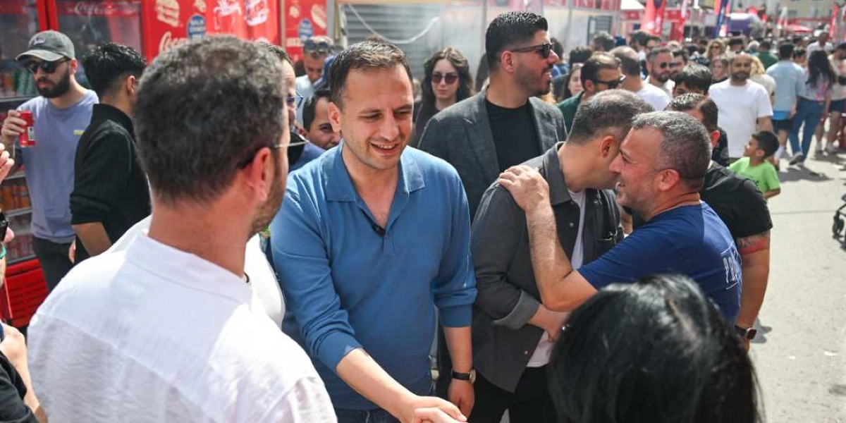 Açık havada düzenlenen bir festival alanında, mavi polo yaka tişörtlü bir erkek, beyaz gömlekli bir kişiyle el sıkışırken gülümsüyor. Çevresinde kalabalık bir insan grubu bulunan bu kişinin hemen yanında, gri ceketli bir adam bir başkasının omzuna elini koyarak sohbet ediyor. Arka planda kırmızı renkli içecek dolapları ve festival alanındaki stantlar görülüyor.