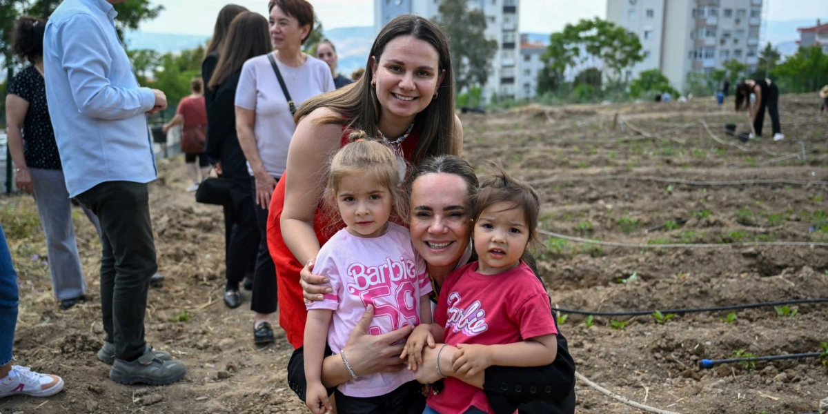 Açık havada, toprak bir alanda iki kadın, kucaklarında küçük çocuklarla birlikte gülümseyerek poz veriyor. Çocuklardan biri üzerinde 