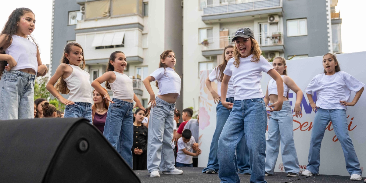 Beyaz tişört ve açık renkli kot pantolon giymiş bir grup kız çocuğu, dış mekanda kurulan bir sahnede dans gösterisi sergiliyor. Ön planda, üzerinde şapka bulunan bir çocuk neşeyle şarkı söyleyerek veya dans ederek performansa öncülük ediyor. Arka planda çok katlı apartmanlar ve etkinliğin adının kısmen göründüğü bir pano yer alıyor.