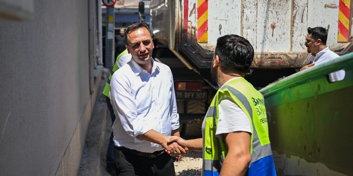Bornova Belediye Başkanı Ömer Eşki, Çamdibi'ndeki bir yol çalışması alanında üzerinde belediye yeleği bulunan bir işçiyle tokalaşıyor. Arka planda bir damperli kamyon ve çalışma yapan diğer görevliler yer alıyor.