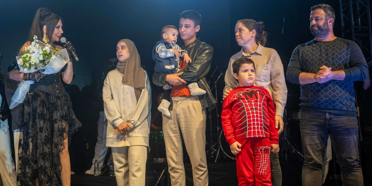 Siyah dantel detaylı bir elbise giymiş, elinde mikrofon ve çiçek buketi tutan bir kadın sanatçı, sahnede bir aile ile birlikte duruyor. Ailede başörtülü genç bir kadın, kucağında bebek taşıyan genç bir erkek, Örümcek Adam desenli kırmızı bir kıyafet giyen küçük bir çocuk, bir kadın ve sakallı bir erkek yer alıyor. Hepsi sahne ışıkları altında yan yana durarak izleyicilere bakıyor.