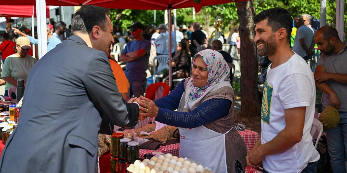 Kırmızı bir çadırın altında kurulan stantta, gri takım elbiseli bir yetkili, yöresel kıyafetli ve başörtülü bir kadınla el sıkışıyor. Kadının yanında beyaz tişörtlü, gülen bir erkek duruyor. Stant masasının üzerinde kavanozlar ve bir tepsi yumurta bulunuyor; arka planda ise etkinliğe katılan diğer insanlar yer alıyor.