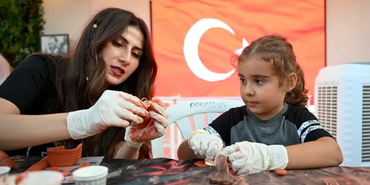 İzmir Enternasyonal Fuarı'ndaki Bornova Belediyesi standında, ellerinde beyaz eldivenlerle kil hamurundan figürler yapan uzun kahverengi saçlı bir kadın ve yanındaki küçük bir kız çocuğu masada oturuyor. Arka planda büyük bir Türk bayrağı görseli yer alıyor.