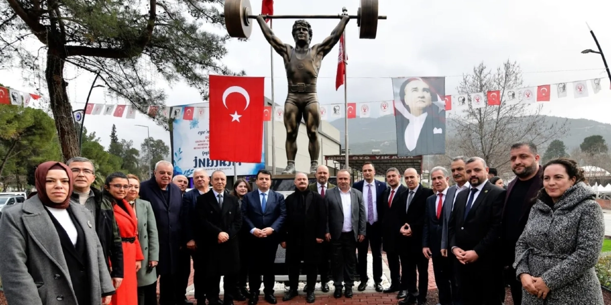 Açık havada, üzerinde halter kaldıran Naim Süleymanoğlu'nun bronz heykelinin bulunduğu bir anıtın önünde poz veren, aralarında Bornova Belediye Başkanı'nın da bulunduğu resmi giyimli bir grup insan. Heykelin arkasında Türk bayrağı ve Mustafa Kemal Atatürk'ün portresinin yer aldığı bir afiş asılıdır; çevrede ise süsleme bayrakları ve ağaçlar görülmektedir.