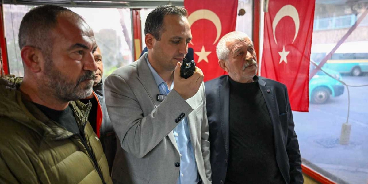 İç mekanda yan yana duran üç erkekten ortadaki kişi, elindeki telsizle konuşurken diğer iki kişi dikkatle onu dinliyor. Ortadaki kişi açık renkli bir ceket ve mavi gömlek giymiş, solundaki kişi haki renkli şişme montlu, sağındaki kişi ise koyu renkli bir ceket ve siyah balıkçı yaka kazak giymiş. Arka planda camın önünde asılı iki adet Türk bayrağı bulunuyor, camın dışından ise bir araç ve sokak manzarası görünüyor.
