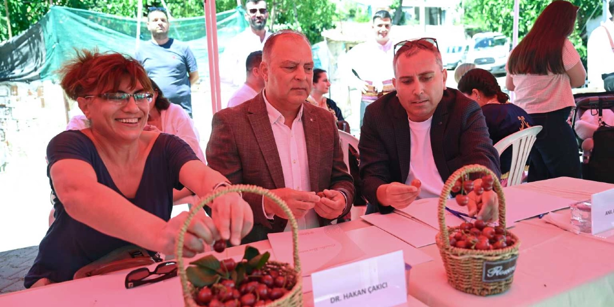 Açık havada kurulan bir masada oturan üç kişi, önlerinde duran hasır sepetlerdeki kirazları inceliyor. Solda gülen bir kadın, ortada ceketli bir erkek ve sağda beyaz tişört üzerine ceket giymiş bir erkek yer alıyor. Masanın üzerinde 