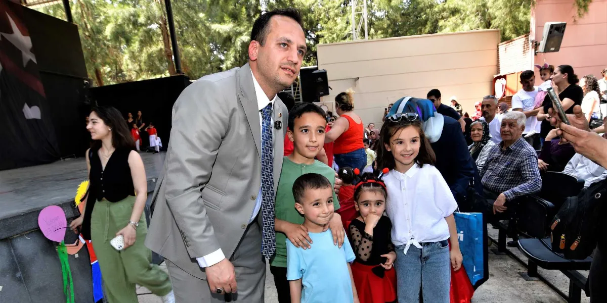 Gri takım elbiseli bir erkek, açık hava etkinlik alanında etrafındaki çocuklarla birlikte gülümsüyor. Çevredeki izleyiciler arasında aileler ve çocuklar bulunurken, arka planda ağaçlar ve sahne düzeni yer alıyor.