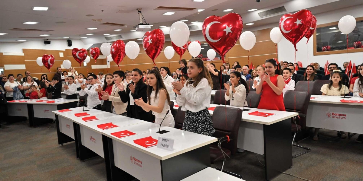 Bornova Belediyesi meclis salonunda, 23 Nisan Ulusal Egemenlik ve Çocuk Bayramı kutlamaları kapsamında sıralarda oturan çocuklar alkışlıyor. Salon, üzerinde Türk bayrağı baskısı bulunan kırmızı kalp şeklinde balonlar ve beyaz balonlarla süslenmiş; masaların üzerinde ise küçük Türk bayrakları ve 