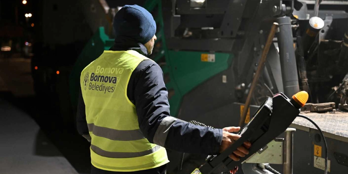 Gece vakti Bornova Belediyesi logolu sarı reflektörlü yelek giyen bir işçi, asfalt serme makinesini elindeki kumanda paneliyle yönetiyor.