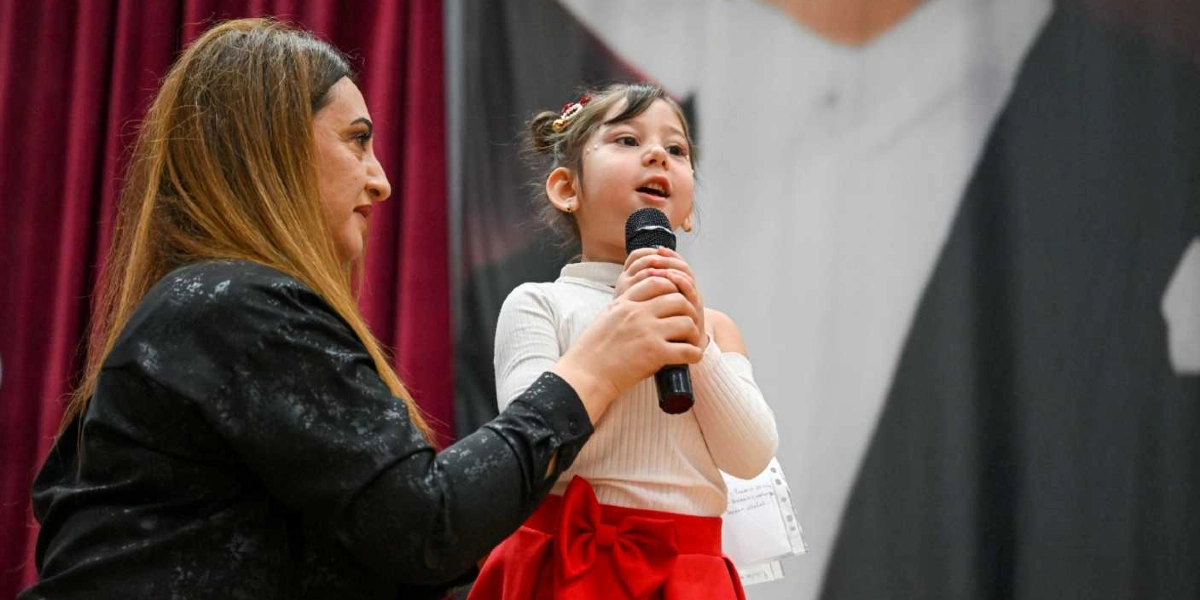 Sahne üzerinde elinde mikrofon tutan küçük bir kız çocuğu, yanında duran bir yetişkinin desteğiyle konuşma yapıyor. Kız çocuğu beyaz bir bluz ve kırmızı fiyonklu bir etek giymiş, saçları toplanmış durumda. Yanındaki kadın ise siyah bir ceket giyiyor ve kızın mikrofonu tutmasına yardımcı oluyor. Arka planda bordo renkli perdeler ve Mustafa Kemal Atatürk'ün silüetinin yer aldığı bir afiş bulunuyor.