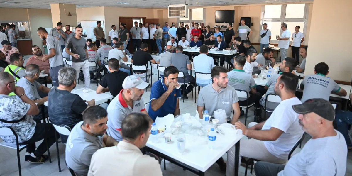 Bornova Belediyesi Park ve Bahçeler Müdürlüğü personelinin bulunduğu kalabalık bir yemekhanede, masalarda oturan ve ayakta duran çok sayıda çalışan bir arada yemek yiyor. Bazı çalışanlar üzerinde Bornova Belediyesi logolu gri tişörtler giyerken, ortamda samimi bir sohbet havası hakim.