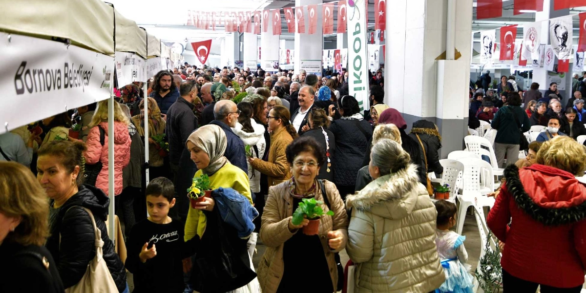 Kapalı bir alanda düzenlenen Tohum Takas Şenliği'nde, Bornova Belediyesi logolu stantların önünde toplanmış kalabalık bir insan grubu görülüyor. Katılımcılar ellerinde saksılarda küçük bitkiler ve tohum paketleri tutuyor, birbirleriyle sohbet ediyor. Tavandan sarkan Türk bayrakları ve çeşitli kurum flamalarıyla süslenmiş alanda, her yaştan insan tohum paylaşımı için bir arada bulunuyor.