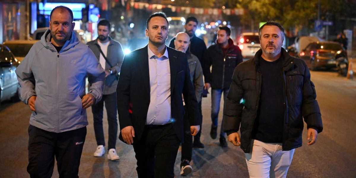 Gece vakti Bornova sokaklarında yürüyen, aralarında takım elbiseli ve günlük kıyafetli kişilerin bulunduğu altı kişilik bir grup. Önde, ortada lacivert ceketli ve açık renk gömlekli bir kişi, yanında ise spor ceketli ve montlu kişiler yer alıyor. Arka planda park halindeki araçlar ve şehir ışıkları görülüyor.