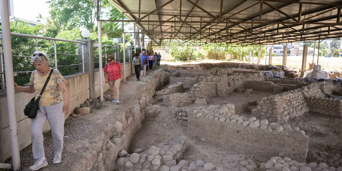 Üstü metal bir çatı ile kapatılmış açık hava arkeolojik kazı alanında yürüyen bir grup insan. Önde sarı çizgili tişörtlü ve gözlüklü bir kadın, arkasında kırmızı puantiyeli bluz giyen başka bir kadın ve onları takip eden bir grup ziyaretçi, taş duvar kalıntılarının yanındaki toprak yolda ilerliyor.