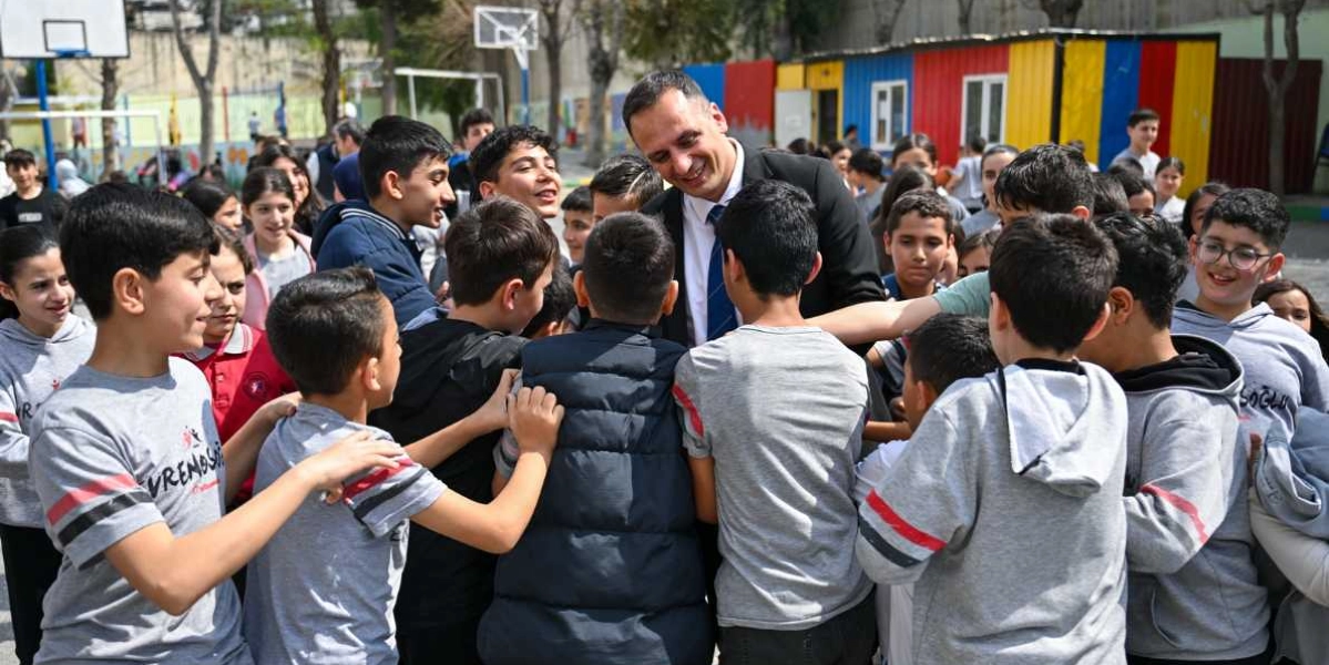 Koyu renk takım elbiseli ve kravatlı bir erkek, okul bahçesinde etrafını saran çok sayıda öğrenciyle bir arada gülümseyerek sohbet ediyor. Öğrencilerin çoğu gri tişörtler giyiyor ve birçoğu adamın etrafında toplanmış, ilgiyle ona bakıyor. Arka planda basketbol potası ve renkli konteyner sınıflar görülüyor.