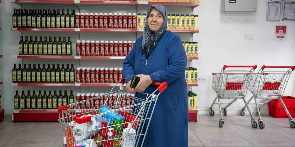 Başörtülü yaşlı bir kadın, market reyonlarının önünde içinde temel gıda ve temizlik ürünleri bulunan alışveriş arabasına tutunarak kameraya bakıyor. Arka planda raflarda düzenli bir şekilde dizilmiş zeytinyağı, ketçap ve mayonez şişeleri görülüyor.