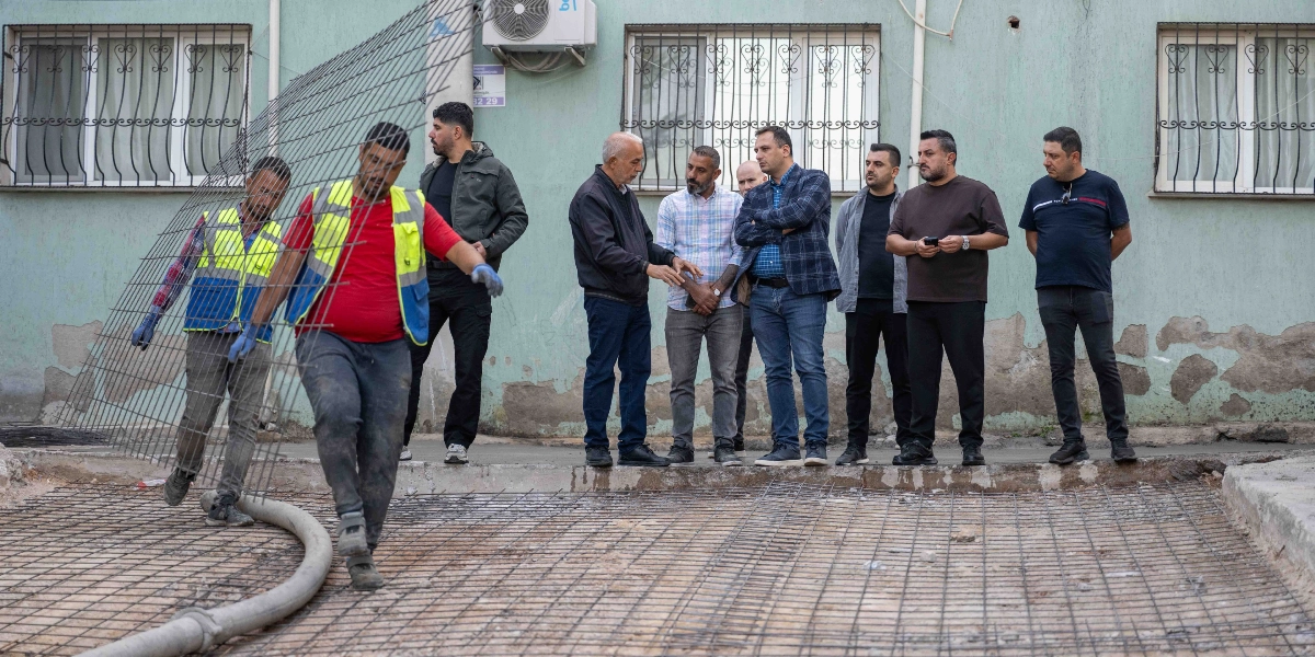 İki inşaat işçisi, yol çalışması için hazırlanan demir donatı ağını yerleştirirken, yanlarında duran bir grup erkek çalışmaları izliyor. İşçilerden biri üzerinde sarı reflektörlü yelek ve kırmızı tişörtle demir ağı tutarken, diğeri mavi yeleğiyle ona yardımcı oluyor. Arka planda ise açık yeşil boyalı, pencerelerinde demir parmaklıklar bulunan bir apartman binası yer alıyor.
