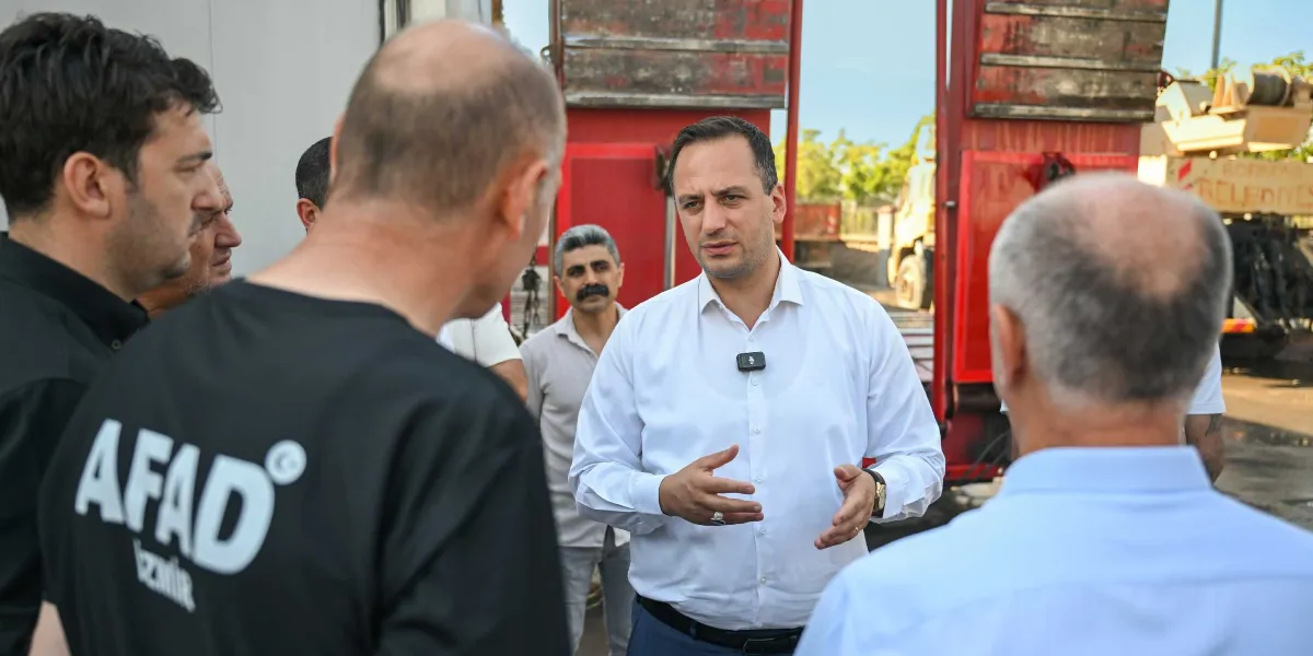 Beyaz gömlekli bir yetkili, üzerinde AFAD İzmir yazılı siyah tişört giyen bir görevli ve diğer yetkililerle bir arada durarak konuşma yapıyor. Arka planda kırmızı renkli bir iş makinesi ve Bornova Belediyesi yazılı bir araç parçası görülüyor.