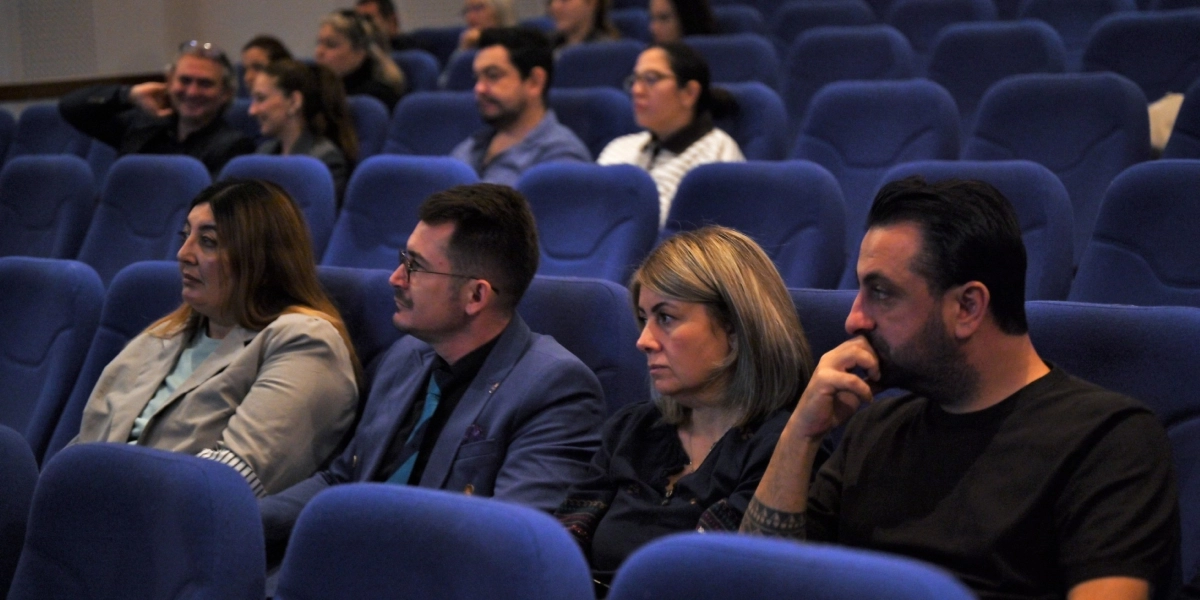 Konferans salonunda mavi koltuklarda oturan ve dikkatle sahneye bakan bir grup insan. Ön sırada soldan sağa doğru açık renk ceketli bir kadın, mavi ceketli ve gözlüklü bir erkek, koyu renk bluzlu bir kadın ve siyah tişörtlü, sakallı bir erkek yer alıyor. Arka sıralarda da etkinliği takip eden diğer katılımcılar bulunuyor.