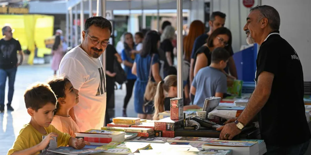 Açık havada kurulan bir kitap fuarında, bir stant görevlisi ile kitapları inceleyen bir baba ve iki çocuğu birbirine gülümseyerek bakıyor. Önlerinde kitaplarla dolu bir stant masası bulunurken, arka planda fuarı gezen diğer insanlar ve stantlar yer alıyor.
