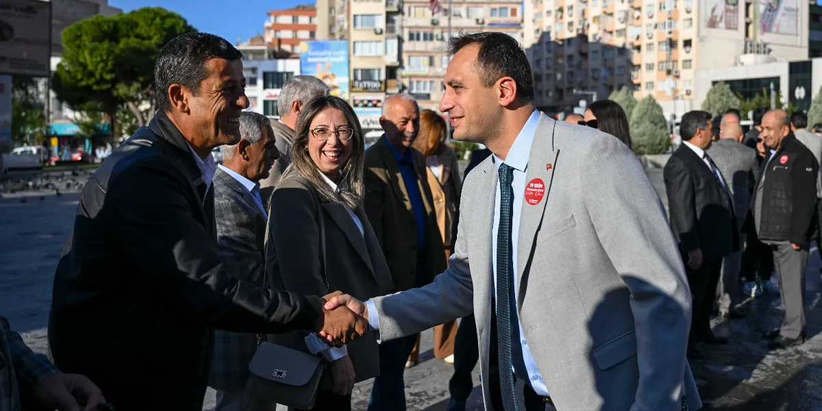 Açık havada, şehir meydanında gri ceketli ve kravatlı bir erkek, karşısındaki kişiyle tokalaşırken gülümseyerek selamlaşıyor. Ceketinin yakasında üzerinde 