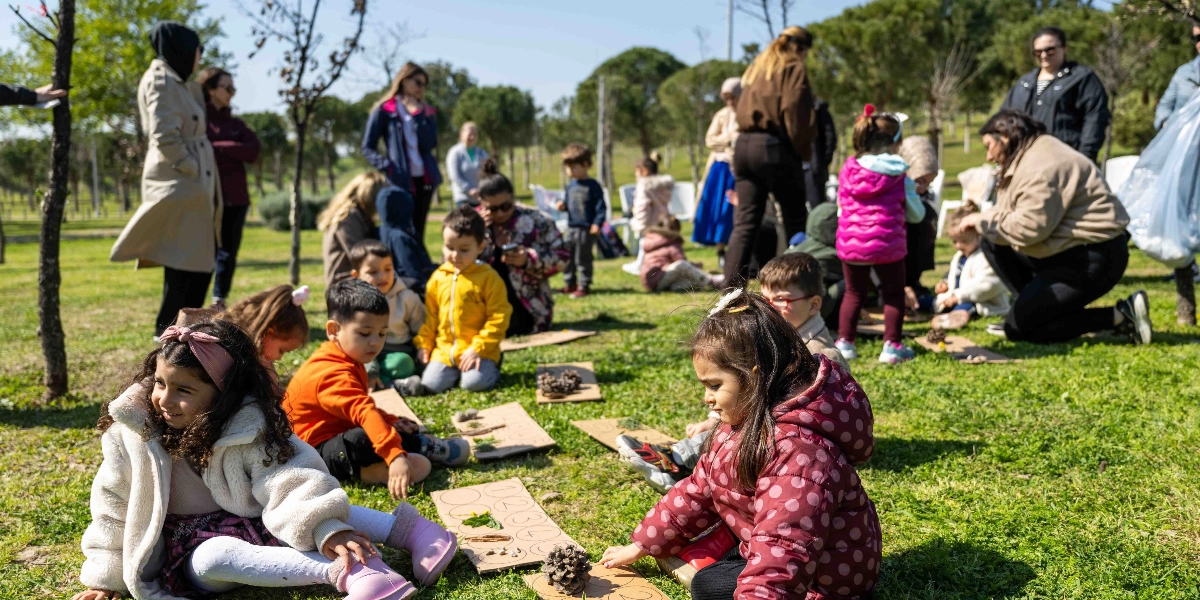 Açık havada, yeşil bir alanda toplanmış çocuklar ve onlara eşlik eden yetişkinler, yere serdikleri kartonlar üzerinde doğadan topladıkları kozalak, dal ve yaprak gibi materyallerle oyunlar oynuyor. Ön planda, beyaz kürklü mont giyen kıvırcık saçlı bir kız çocuğu ile bordo puantiyeli mont giyen bir başka kız çocuğu, kartonların üzerindeki sayıları ve nesneleri inceliyor. Arka planda ise sarı montlu bir çocuk ve diğer arkadaşları kendi kartonlarıyla ilgilenirken, yetişkinler çocukları gözlemliyor. Güneşli bir günde çekilen bu kare, doğa temalı bir eğitim etkinliğini yansıtıyor.