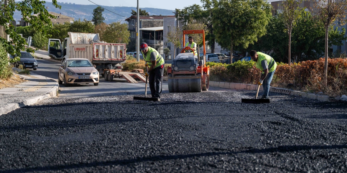 Bornova Belediyesi ekipleri, bir cadde üzerinde asfalt serme ve düzeltme çalışması yapıyor. Sarı reflektörlü yelek giyen işçiler, ellerindeki tırmıklarla yeni dökülen sıcak asfaltı düzeltirken, arkalarında turuncu renkli bir silindir aracı ilerliyor. Arka planda ise üzerinde Bornova Belediyesi yazan bir damperli kamyon ve trafikte bekleyen araçlar görülüyor.