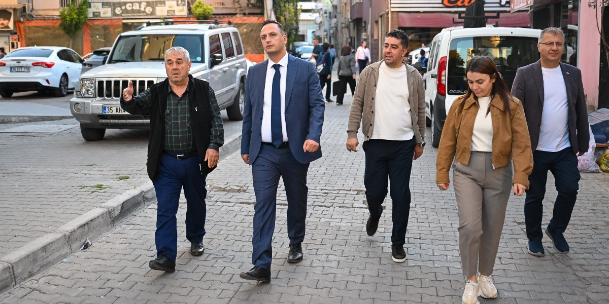 Kilit taşı döşeli bir sokakta yürüyen beş kişilik grup; en solda elini ileriye doğru uzatarak bir şeyler anlatan yaşlı bir adam, yanında lacivert takım elbiseli ve kravatlı bir erkek, onun yanında bej hırka ve beyaz tişört giyen bir erkek, yanında kahverengi ceketli bir kadın ve en sağda gri ceketli bir erkek yer alıyor.