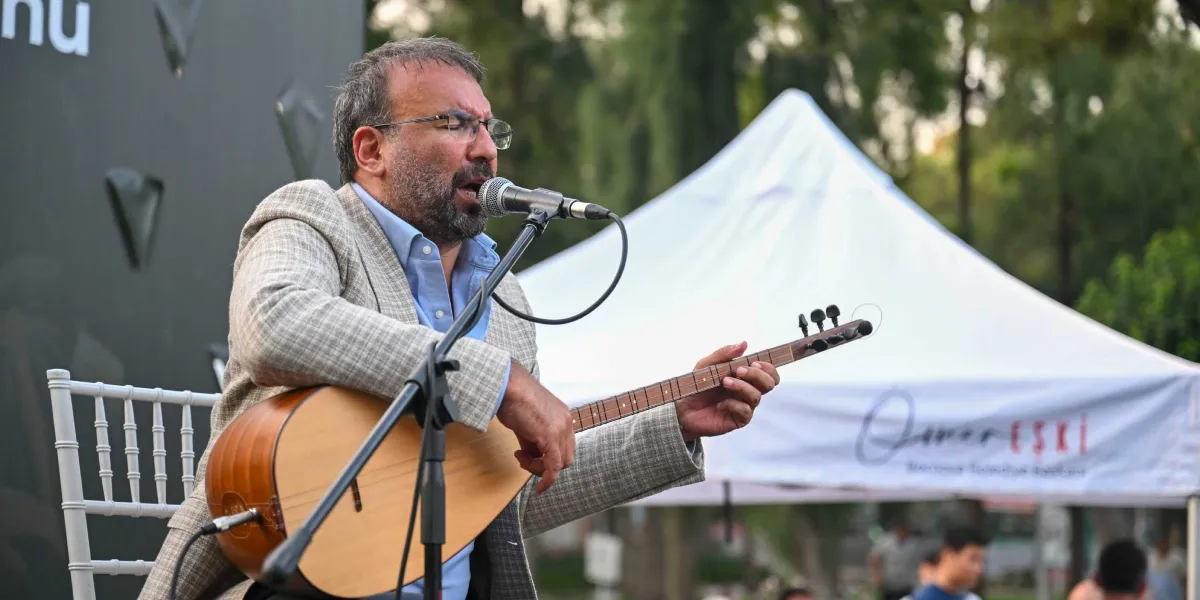Gözlüklü, sakallı, açık renkli ekose bir ceket ve mavi gömlek giymiş bir adam, açık havada elinde bağlama çalarak mikrofon başında şarkı söylüyor. Arka planda üzerinde 