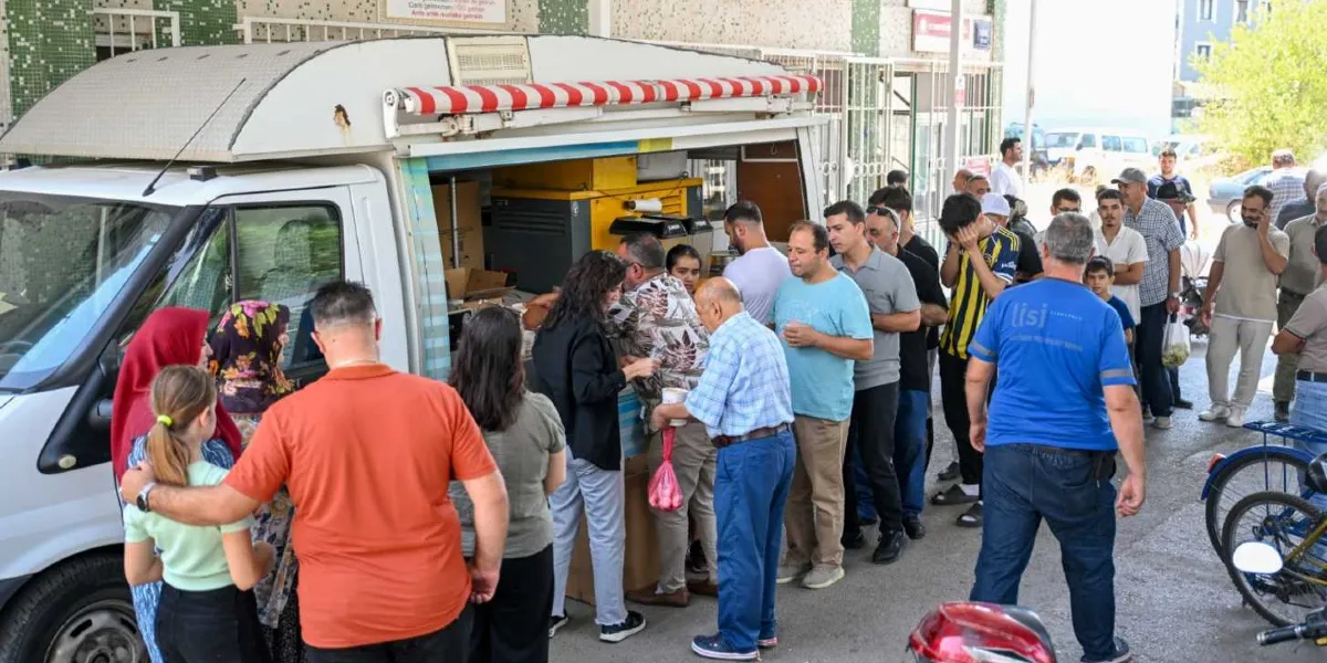 Bornova Belediyesi'ne ait beyaz bir mobil ikram aracının önünde, lokma veya benzeri bir yiyecek dağıtımı için sıra bekleyen kalabalık bir grup insan görülüyor. İnsanlar aracın servis penceresine doğru yönelmiş durumda ve bazıları ellerinde yiyecek paketleriyle bekliyor. Gün ışığında, dış mekanda çekilmiş bu karede farklı yaş gruplarından kadın ve erkekler yer alıyor.