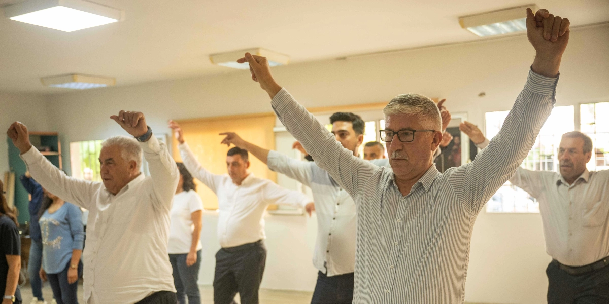 Kapalı bir mekanda yan yana dizilmiş, beyaz gömlekli bir grup erkek ve kadın, ellerini havaya kaldırarak halk oyunları veya benzeri bir etkinlik için figür sergiliyor. Ön planda gözlüklü, çizgili gömlekli bir adam odak noktasında yer alıyor ve diğer katılımcılarla birlikte aynı hareketleri yaparak coşkulu bir şekilde dansa eşlik ediyor.