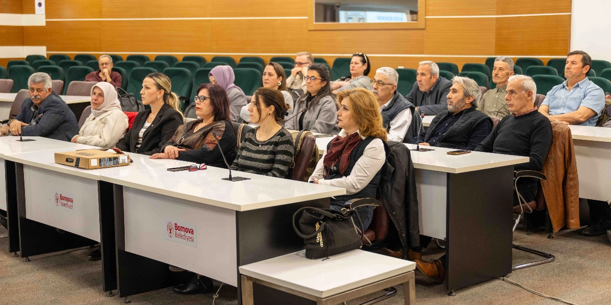 Bornova Belediyesi tarafından düzenlenen dijital dönüşüm eğitimine katılan muhtarlar, konferans salonundaki sıralarda oturarak dikkatle dinliyor. Ön planda beyaz masaların üzerinde Bornova Belediyesi logoları yer alırken, katılımcılar arasında farklı yaş gruplarından kadın ve erkekler bulunuyor. Salonun arka kısmında yeşil renkli koltuklar ve ahşap duvar panelleri görülüyor.