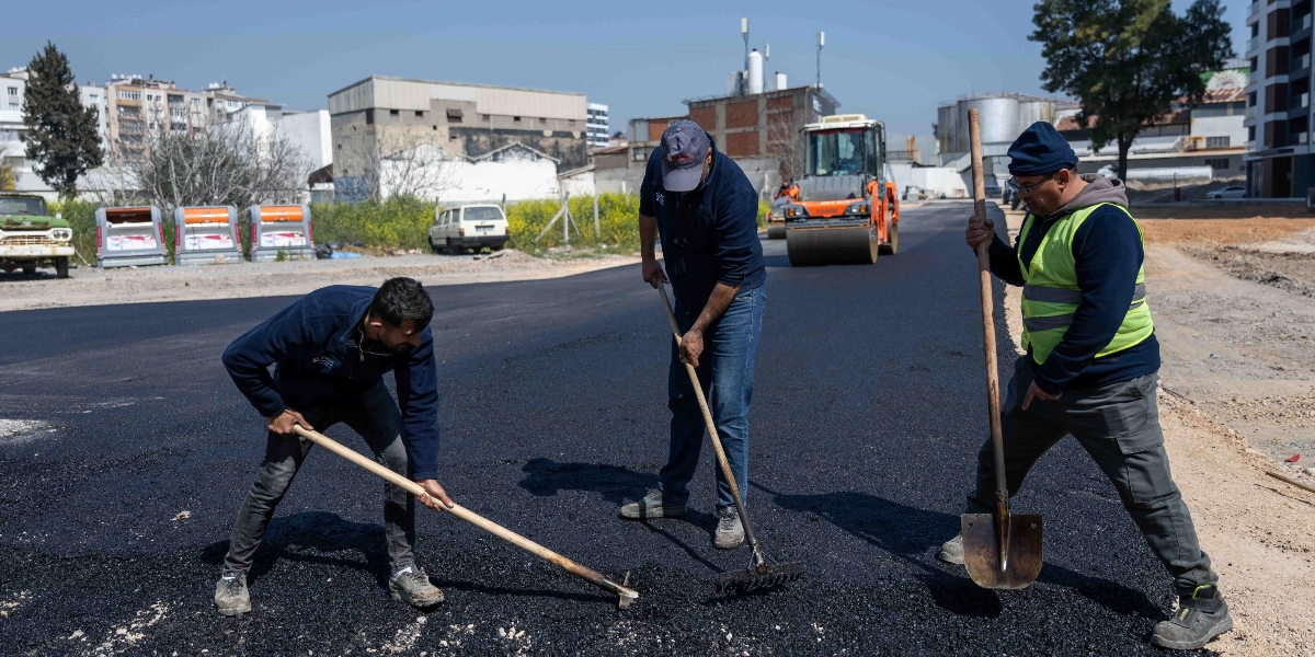 Üç işçi, yeni dökülen asfaltı kürek ve tırmık yardımıyla düzeltiyor. İşçilerden biri sarı reflektörlü yelek giyiyor, diğerleri ise koyu renkli kıyafetlerle çalışıyor. Arka planda yol silindiri ve şehir yerleşimi görülüyor.