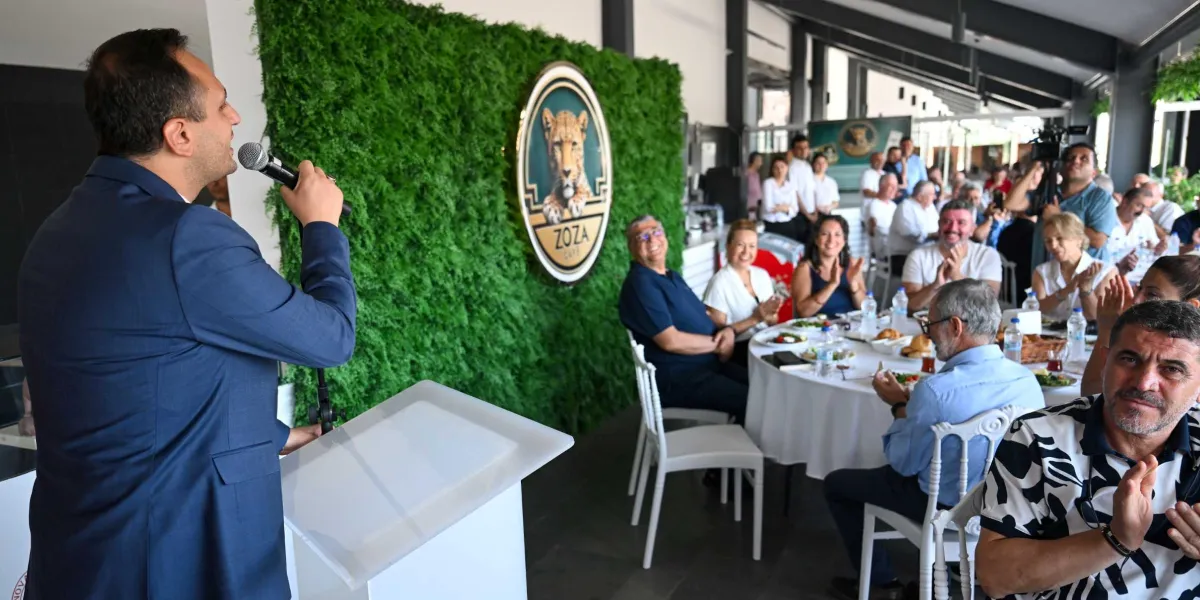 Lacivert ceketli bir konuşmacı, kürsüde mikrofonla hitap ederken, arka planda üzerinde leopar figürü ve Zoza Cafe yazısı bulunan yeşil yapay bitkili bir duvar önünde oturan davetliler kendisini alkışlıyor. Beyaz örtülü masalarda oturan katılımcılar, konuşmayı dikkatle dinleyerek eşlik ediyor.