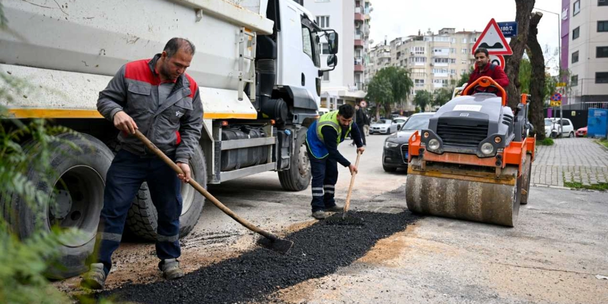 Belediye işçileri, Bornova'da bir cadde üzerinde asfalt yenileme çalışması yapıyor. İşçilerden biri kürekle asfaltı düzeltirken, diğeri tırmıkla çalışıyor ve bir diğeri de silindir makinesini kullanarak asfaltı sıkıştırıyor. Arka planda park halindeki araçlar ve şehir binaları görülüyor.