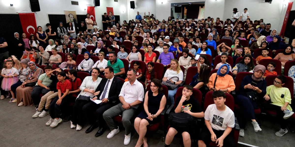Konferans salonunda dolu koltuklarda oturan, aralarında çocukların ve ailelerin bulunduğu kalabalık bir izleyici grubu. Salonun ön sıralarında resmi kıyafetli yetkililer ve katılımcılar yer alırken, arka taraflara doğru uzanan izleyiciler sahneye doğru bakıyor. Salonun duvarlarında Türk bayrakları ve Atatürk posteri asılı bulunuyor.