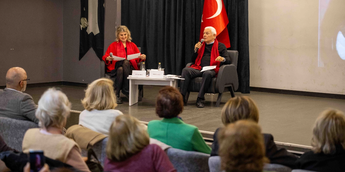 Kapalı bir salonda düzenlenen etkinlikte, sahnede karşılıklı oturan bir kadın ve bir erkek konuşma yapıyor. Kadın kırmızı bir şal takmış, elinde notlar tutuyor; erkek ise siyah kıyafetinin üzerinde kırmızı bir atkı ile mikrofonla konuşuyor. İkilinin önündeki küçük masada isimlikler ve su şişeleri bulunuyor. Arka planda siyah bir perde ve Türk bayrağı yer alıyor. İzleyiciler, sahnedeki konuşmacıları dikkatle dinliyor.