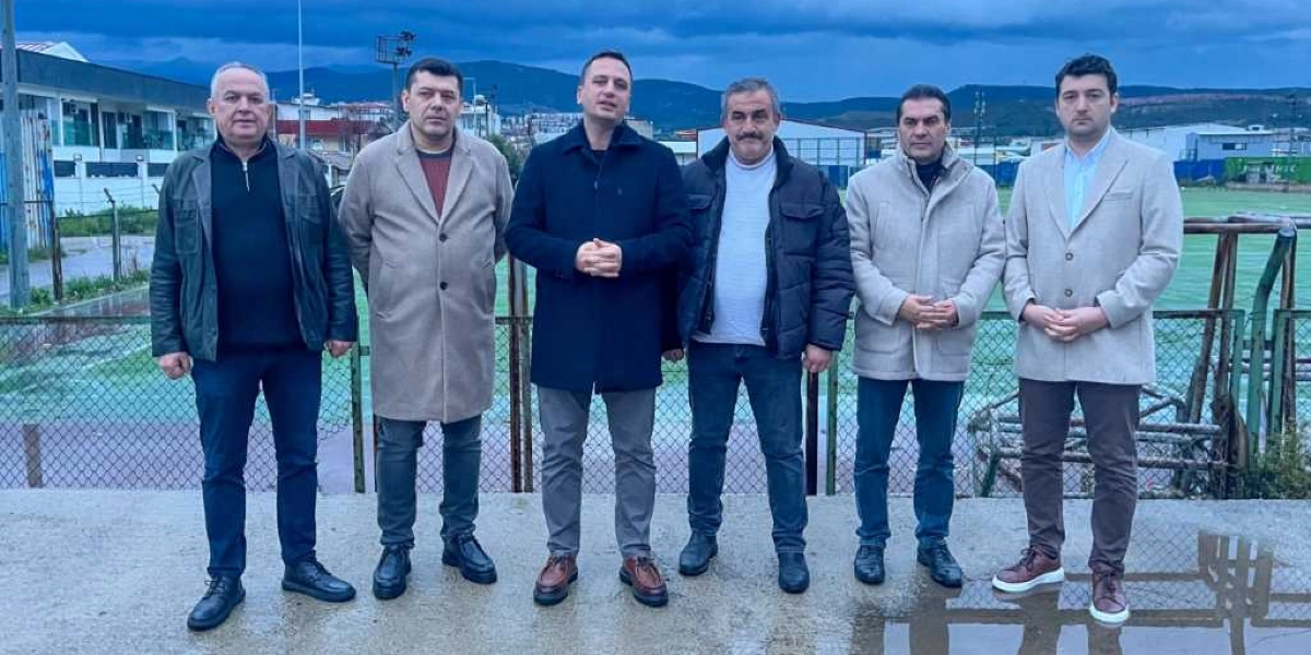 Dış mekanda, arkalarında tel çit ve spor sahası bulunan, kışlık kıyafetler giymiş altı erkek yan yana durarak objektife bakıyor. Ortada yer alan kişi konuşma yapar gibi ellerini birleştirmiş, diğerleri ise elleri önlerinde birleşik veya yanlarında duruyor. Arka planda binalar ve bulutlu bir gökyüzü görülüyor.