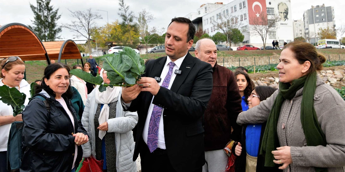 Bornova Kent Bostanları'nda düzenlenen hasat etkinliğinde, takım elbiseli bir yetkili elinde tuttuğu taze brokoliye bakarak çevresindeki vatandaşlara bilgi veriyor. Etkinliğe katılan farklı yaş gruplarından kadınlar ve çocuklar, hasat edilen ürünleri ilgiyle inceliyor. Arka planda üzerinde Türk bayrağı ve Atatürk görseli bulunan bir okul binası ile açık hava bostan alanı görülüyor.