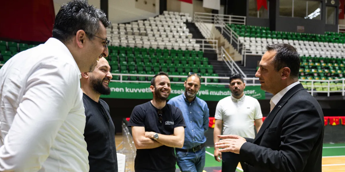 Bornova Belediye Başkanı Ömer Eşki, Göztepe spor tesislerinde bir grup sporcu ve teknik ekiple bir araya gelerek sohbet ediyor. Arka planda yeşil ve beyaz koltuklu tribünlerin yer aldığı kapalı spor salonunda, Başkan Eşki siyah ceketli ve beyaz gömlekli haliyle konuşurken, karşısındaki ekip üyeleri gülümseyerek onu dinliyor.