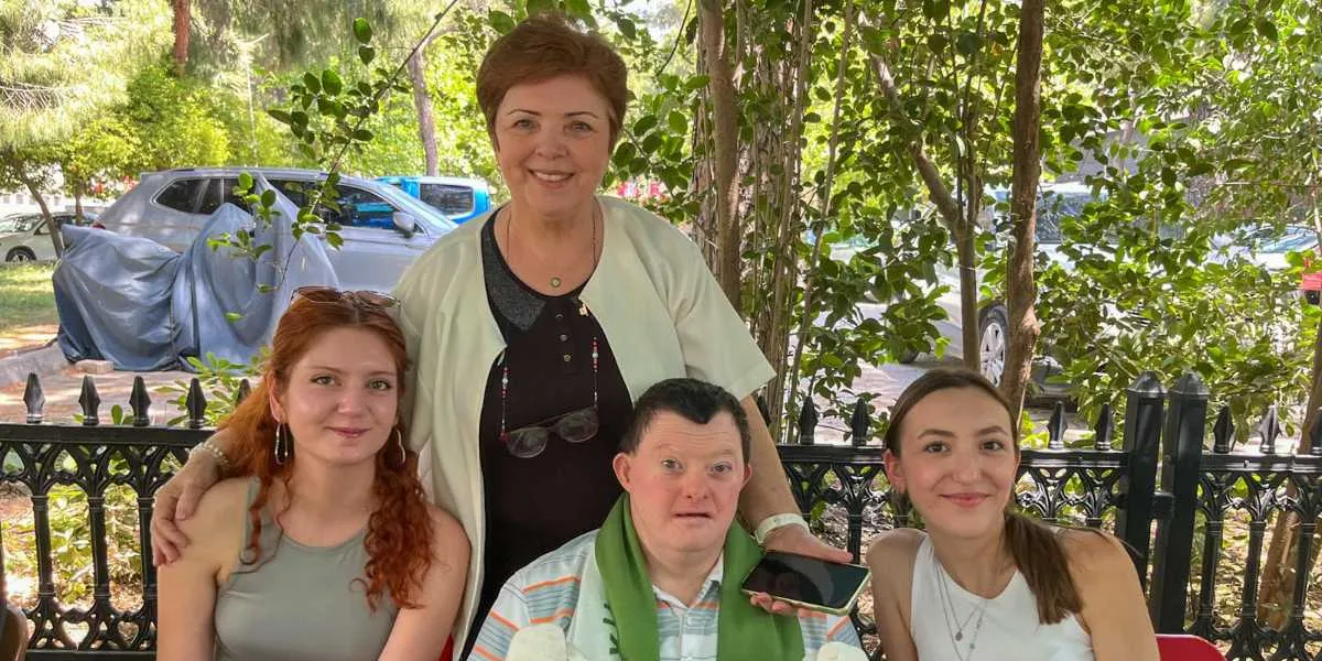 Açık havada, yeşilliklerin ve siyah ferforje bir çitin önünde yan yana duran dört kişi gülümsüyor. Ortada, kısa saçlı, açık renkli bir ceket giyen orta yaşlı bir kadın, bir eliyle yanında oturan Down sendromlu genç bir adamın omzuna dokunuyor. Genç adamın boynunda yeşil bir atkı bulunuyor ve elinde bir cep telefonu tutuyor. Grubun iki yanında ise uzun saçlı, genç iki kadın yer alıyor. Arka planda ağaçlar ve park halindeki araçlar görülüyor.