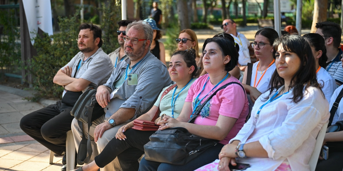 Açık havada, ağaçlık bir alanda sandalyelerde oturmuş, dikkatle bir yöne doğru bakan çeşitli yaş gruplarından oluşan bir grup insan. Katılımcıların birçoğunun boynunda etkinlik kartı asılıdır ve ön planda pembe tişörtlü bir kadın ile beyaz gömlekli bir kadın yer almaktadır. Arka planda ise güneşli bir günde etkinliği takip eden diğer katılımcılar görülmektedir.