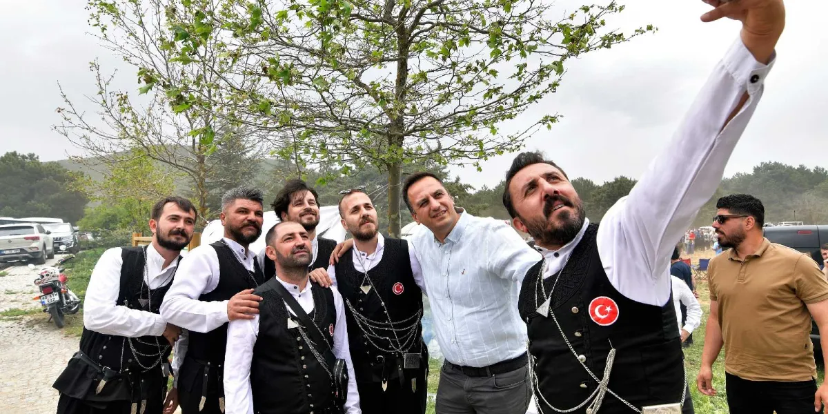 Açık havada, geleneksel Karadeniz kıyafetleri giymiş bir grup erkek ve yanlarında beyaz gömlekli bir beyefendi, hep birlikte özçekim yapıyor. Geleneksel kıyafetli kişiler, üzerinde Türk bayrağı arması bulunan siyah yelekler ve gümüş zincir detaylı aksesuarlar kullanıyor. Grubun en sağında yer alan ve özçekimi yapan kişi, diğerleriyle birlikte gülümseyerek kameraya bakıyor. Arka planda ağaçlar, park halindeki araçlar ve bulanık bir açık alan manzarası görülüyor.